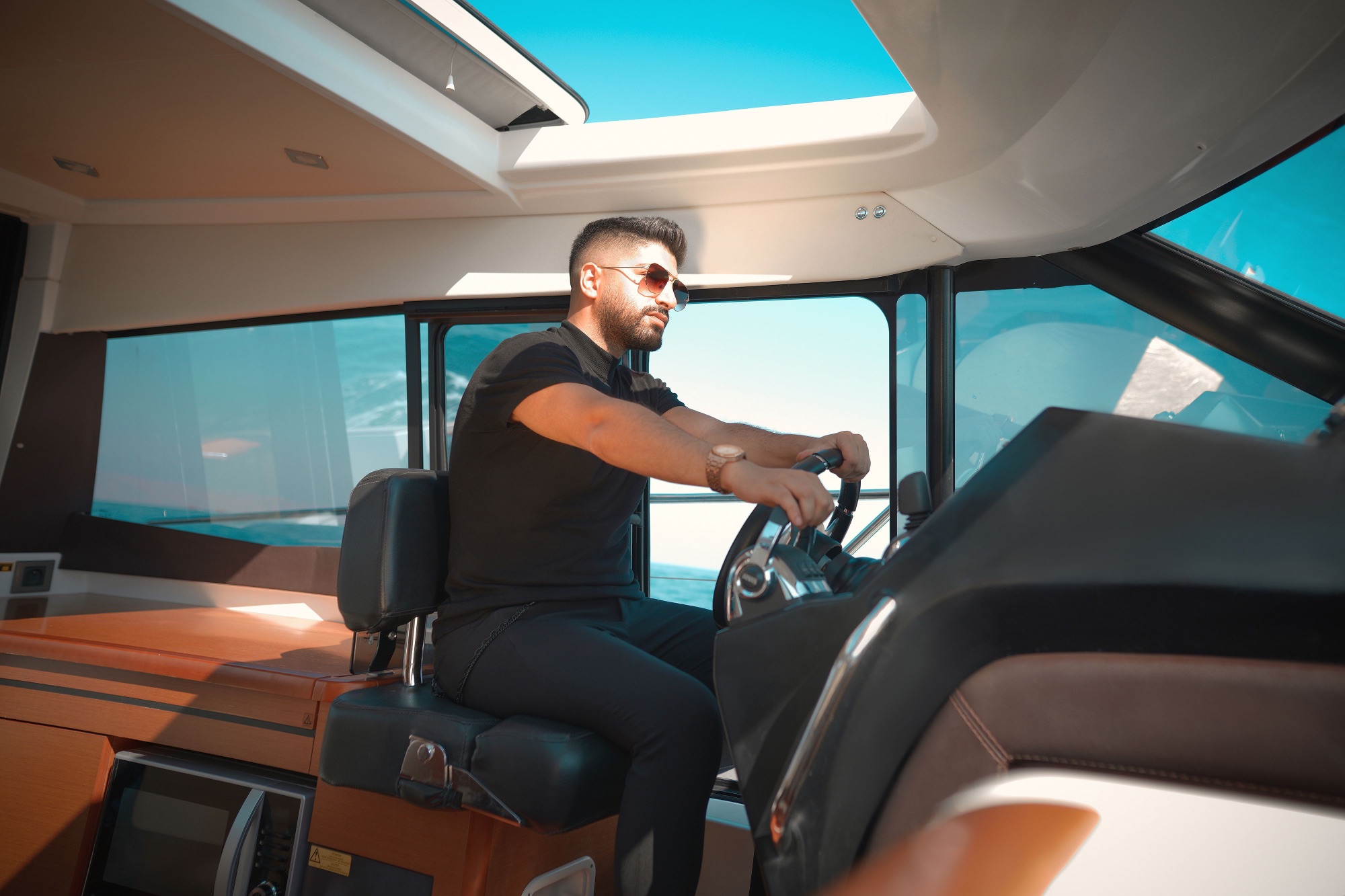 Captain steering from the helm seat of a motor yacht beneath an open sunroof, warm wood and black leather interior