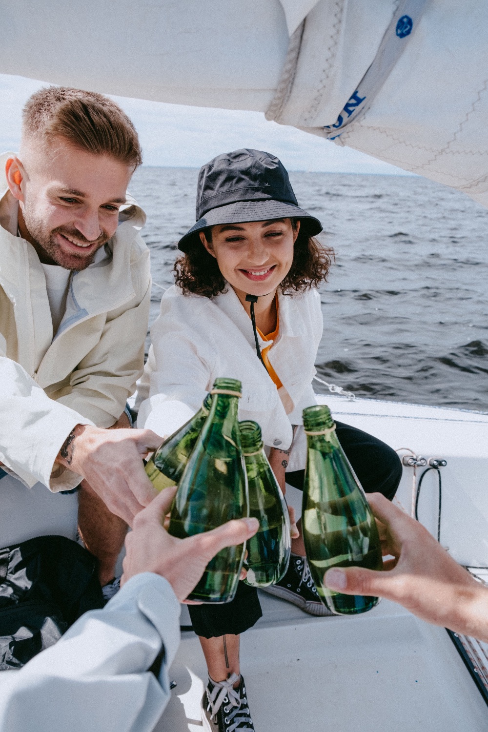 Three friends laughing and toasting with bottles on a sailboat deck beneath the boom and sail