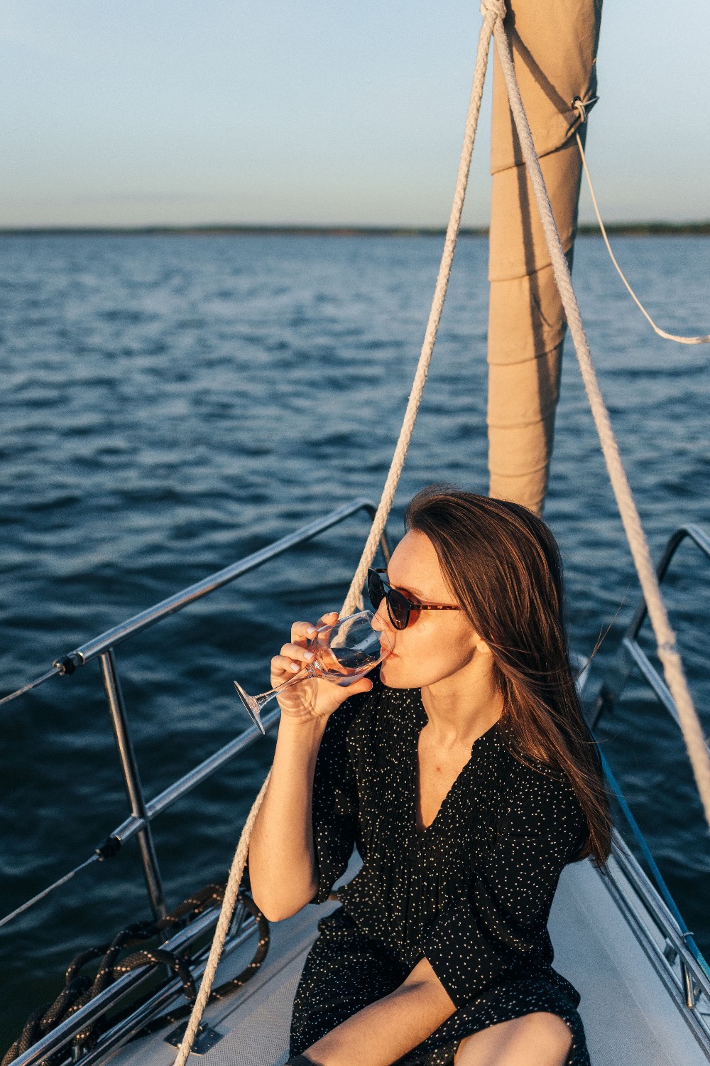 Woman in sunglasses sipping wine at a sailboat's bow in warm evening light over deep blue water